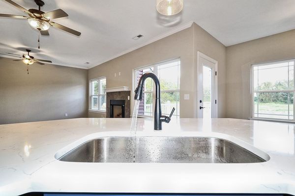 Kitchen sink with running water, in front of a white countertop. Open space with light tan walls, windows, and ceiling fans.
