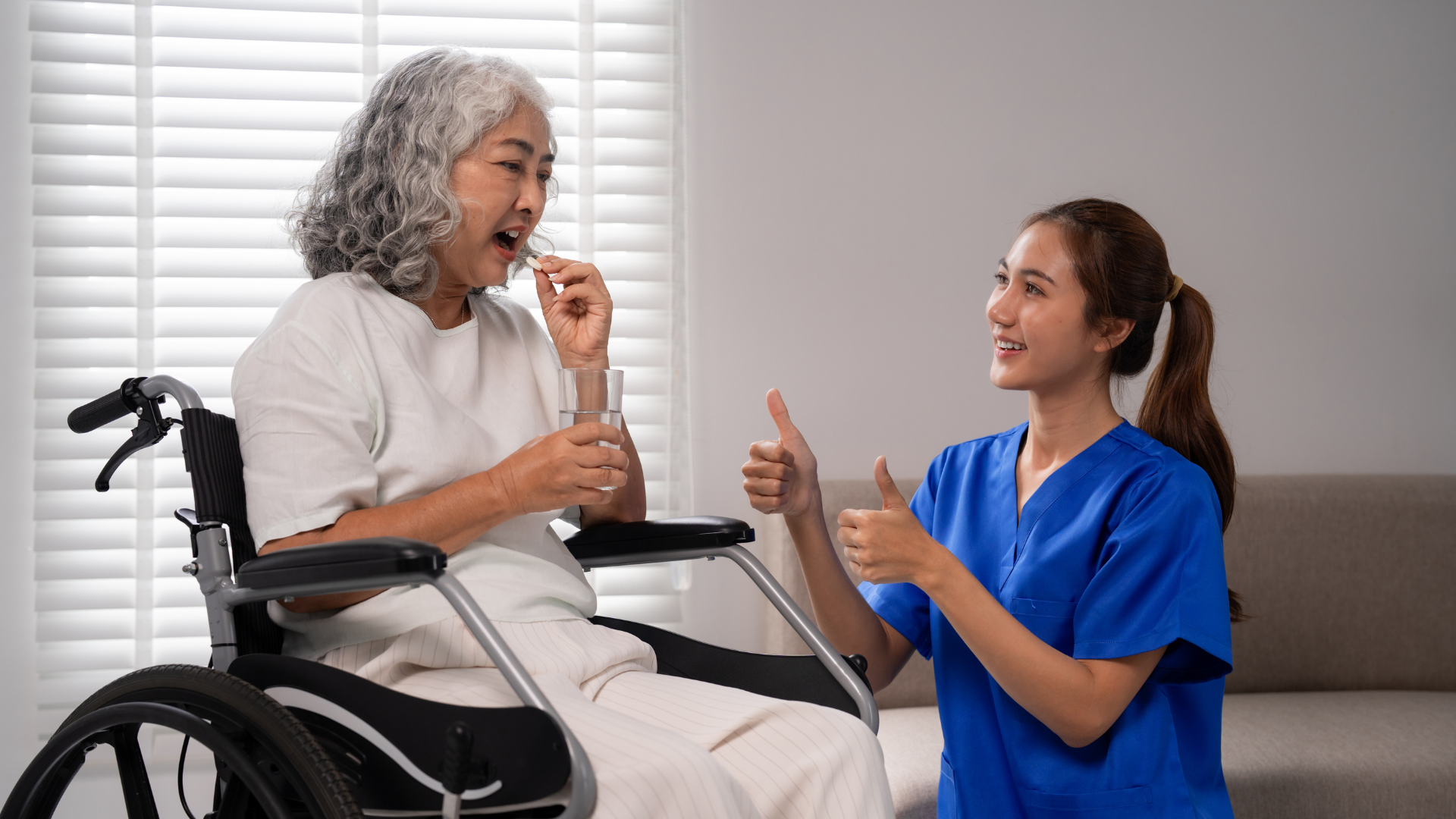 Caregiver in blue scrubs feeding an elderly woman in a wheelchair in a bright room