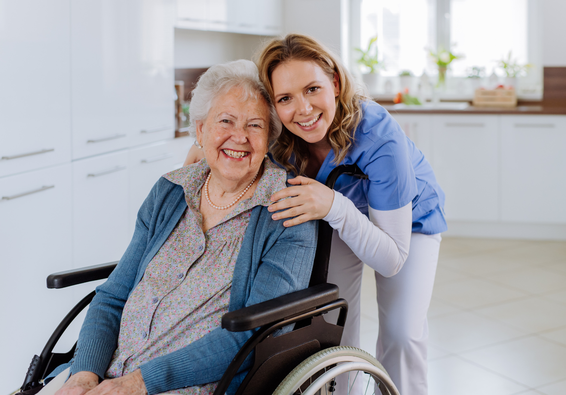 Caregiver in blue uniform smiling and standing behind woman in wheelchair in a bright home kitchen