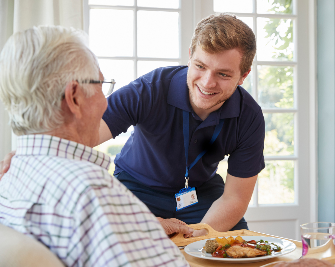 Caregiver smiling and leaning toward a seated man at a dining table, offering support during a meal