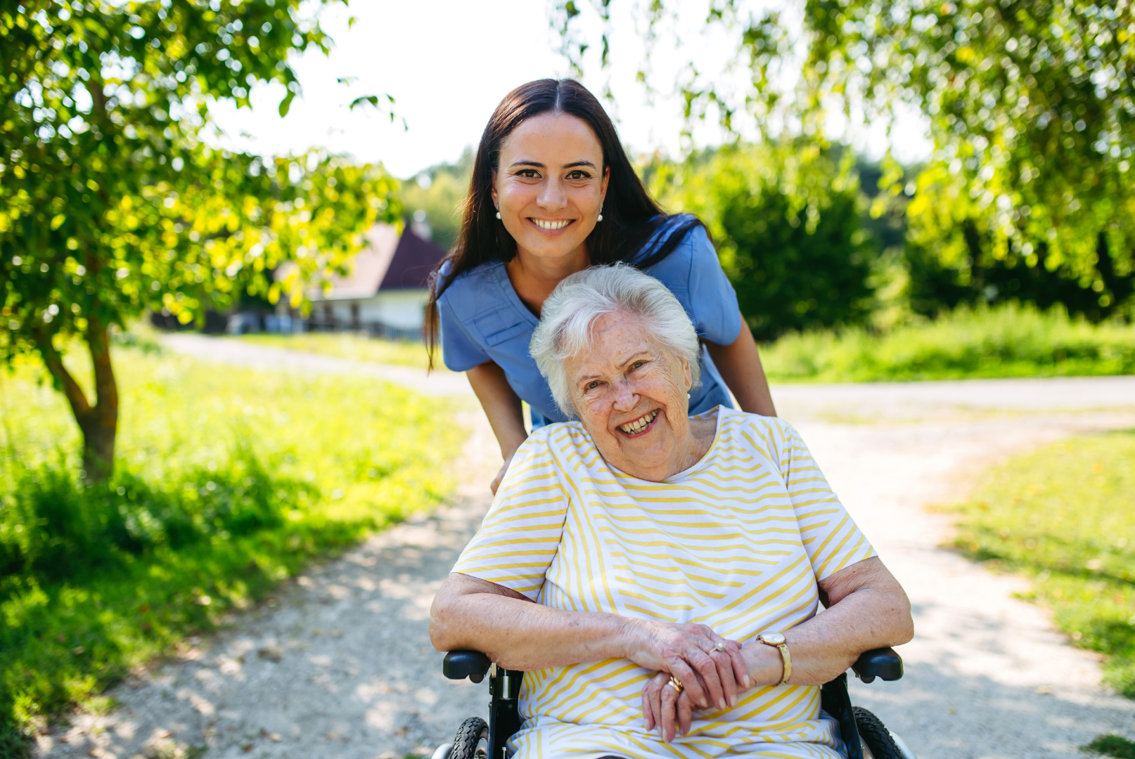 Woman pushes smiling elderly person in wheelchair on a sunlit path outdoors