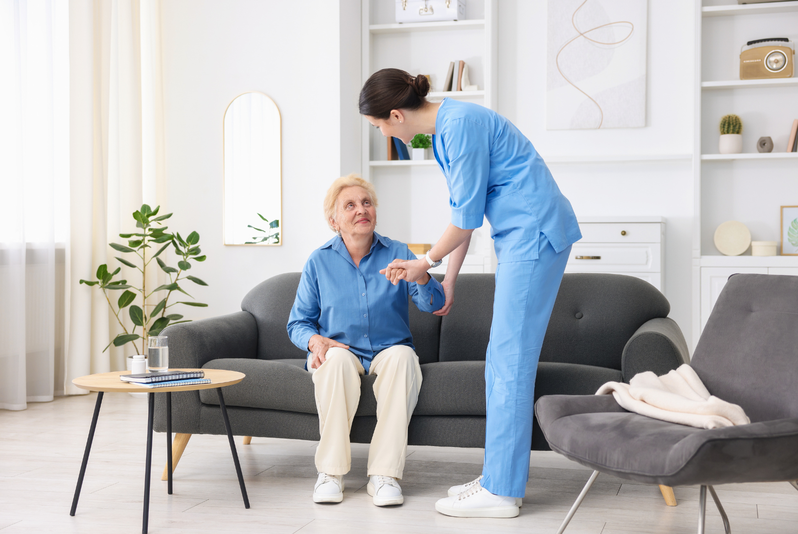Caregiver in blue scrubs helping an elderly man sit on a sofa in a bright living room