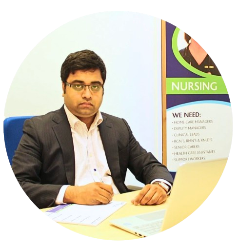 Man in a black suit seated at a desk, writing paperwork beside a nursing recruitment poster.