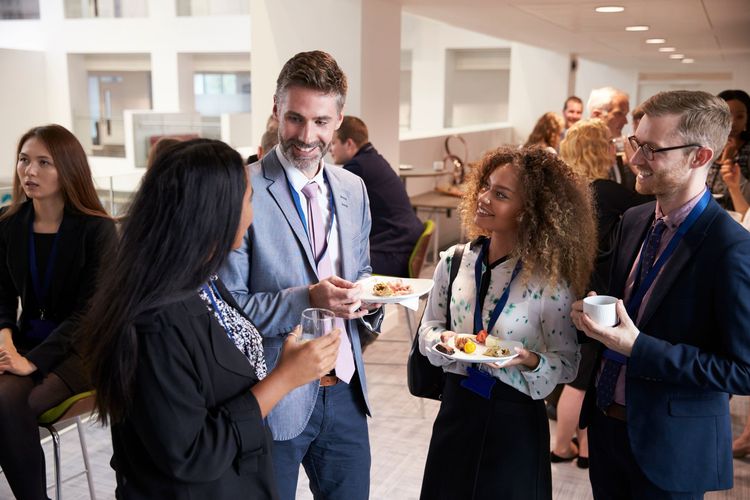 People networking at a conference, holding drinks and food, smiling. Inside a building.