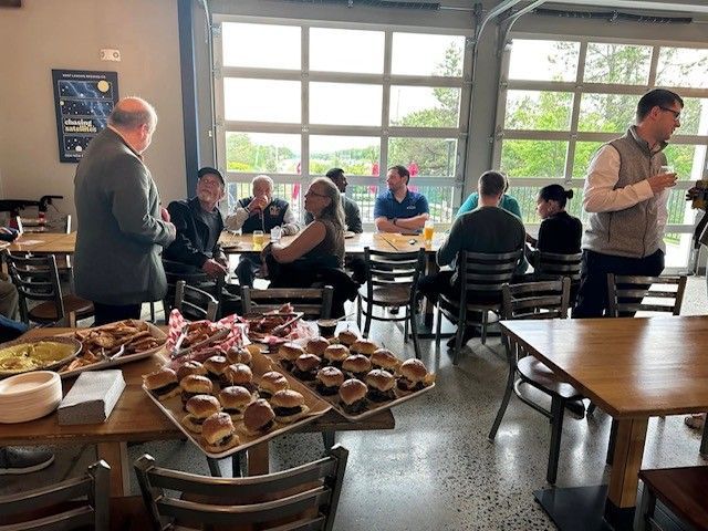 Group of people gathered indoors, eating and socializing around tables with food, near large windows. Group of people gathered indoors, eating and socializing around tables with food, near large windows.