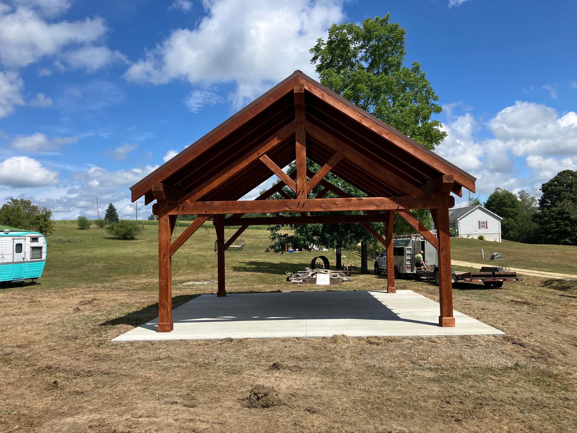 A dark wood timber frame pavilion stands on a concrete pad in a grassy field under a sunny blue sky.