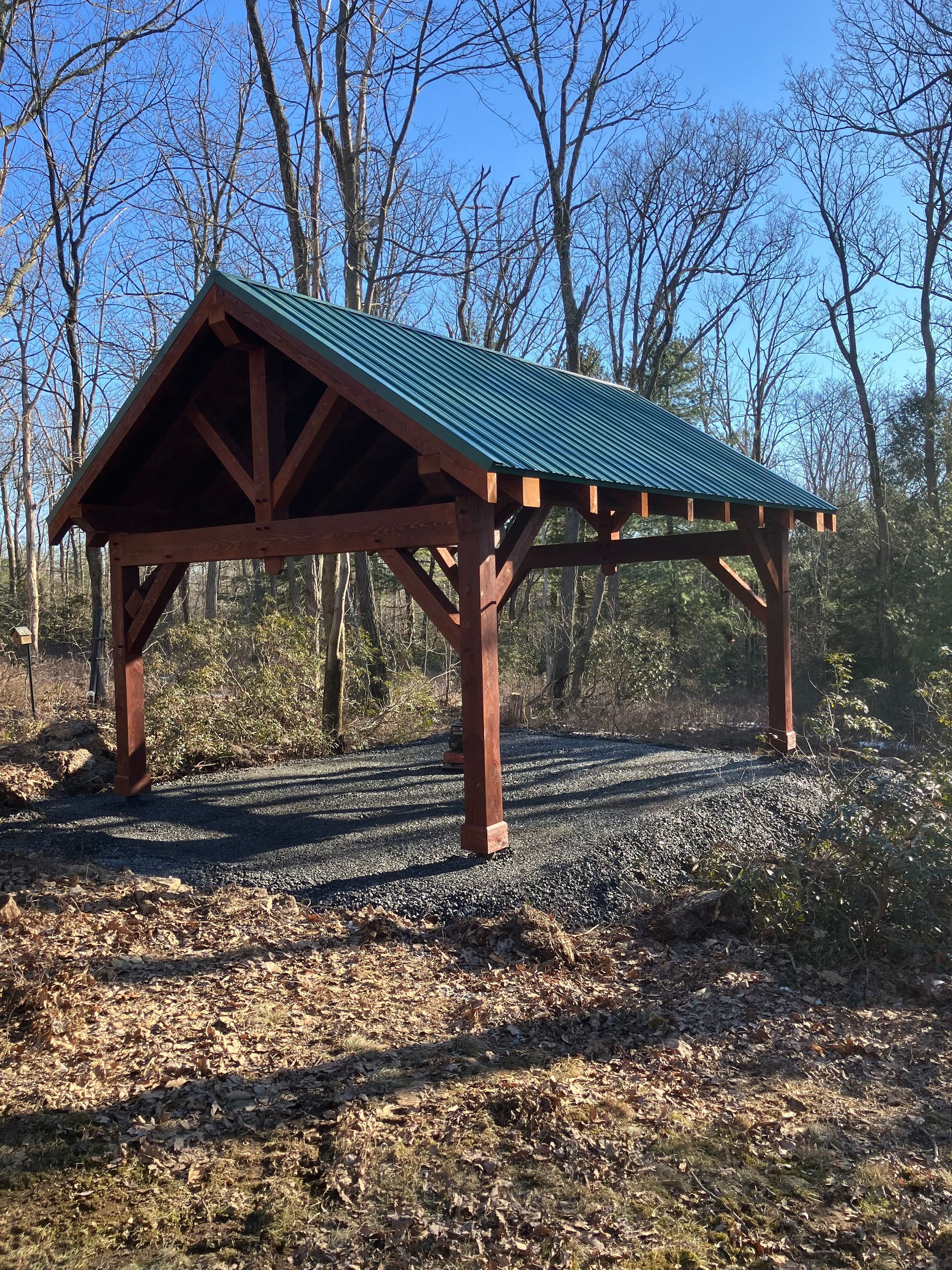 A wooden timber-frame pavilion with a green metal roof stands over a gravel pad in a sunlit forest.