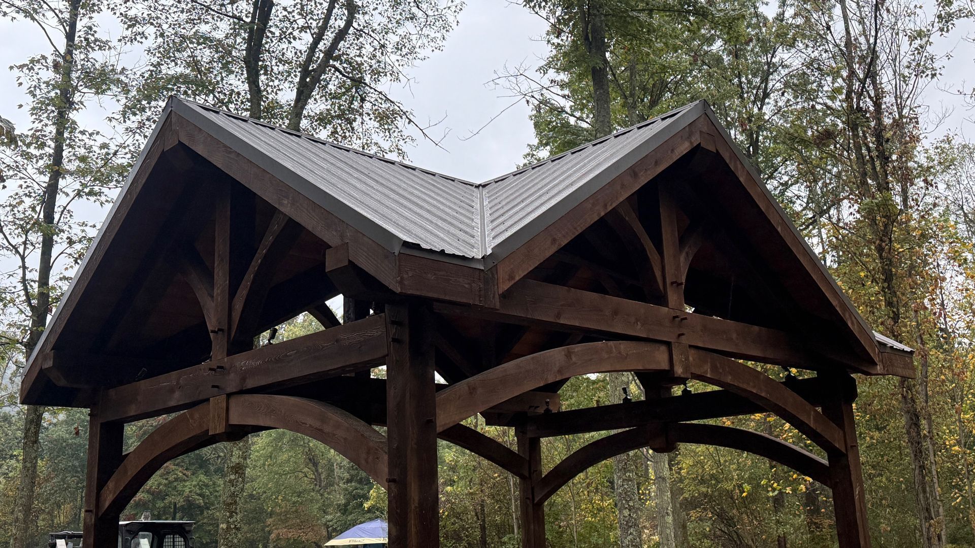 A dark brown wooden pavilion with a peaked, gabled metal roof set against a backdrop of trees.