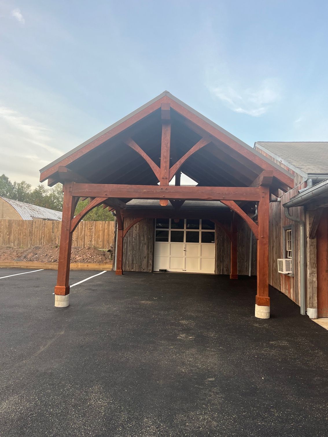 A large, timber-framed wooden carport with a gabled roof standing over an asphalt driveway next to a rustic building.
