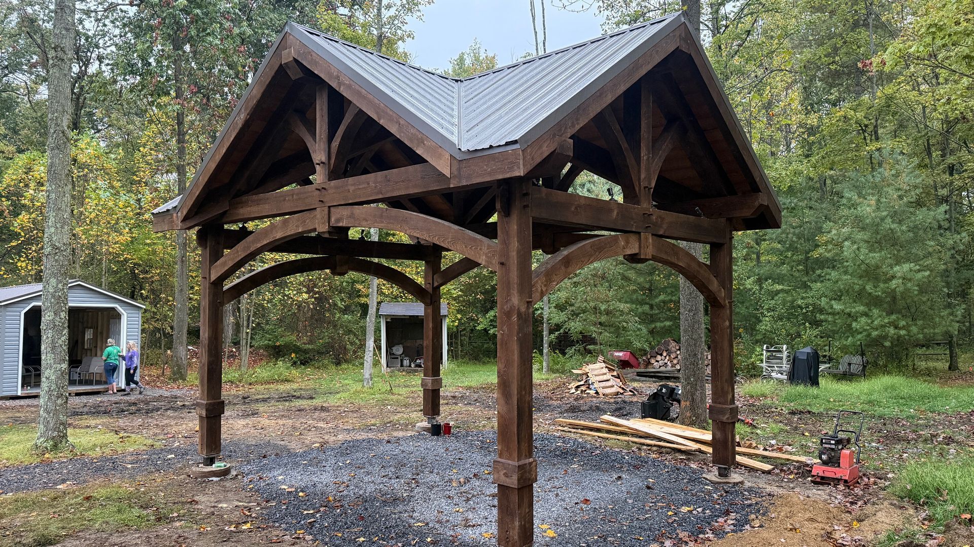 A dark brown, timber-framed pavilion with a double-peaked roof stands on a gravel base in a wooded outdoor setting.
