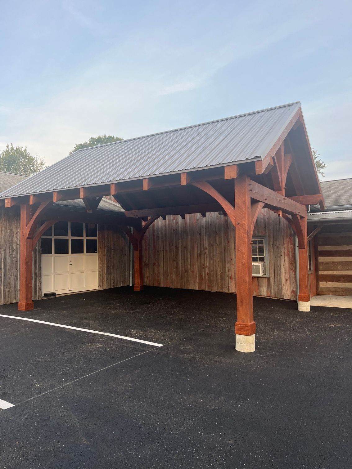 A timber-frame carport with a dark metal roof standing over a black asphalt driveway beside rustic wooden buildings.