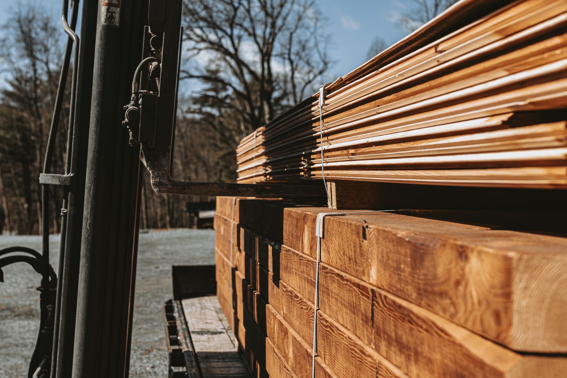 A forklift lifts a large stack of wooden planks and beams outdoors on a sunny day. Lumber by By Central Timber Frame Supply. 