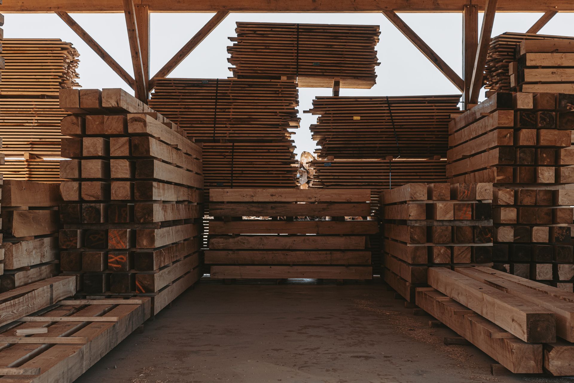 Piles of stacked wooden beams and planks organized inside a large, open-air shed. By Central Timber Frame Supply. 