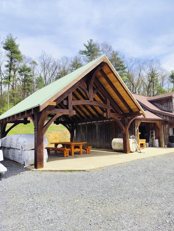 A timber-frame, open-air pavilion with a green metal roof, sheltering a picnic table on a gravel surface outdoors.