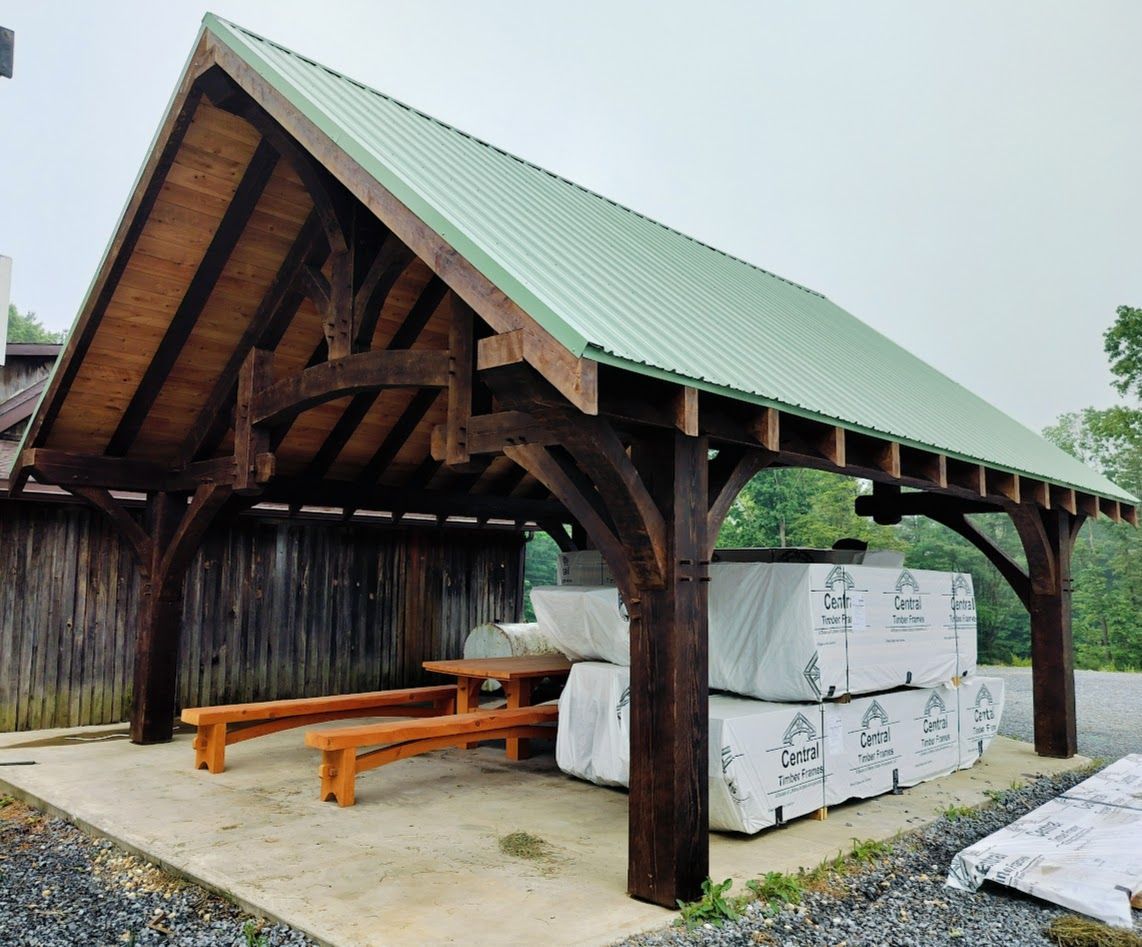 A timber-frame open pavilion with a green metal roof, wood benches, and stacked building materials on a concrete slab.