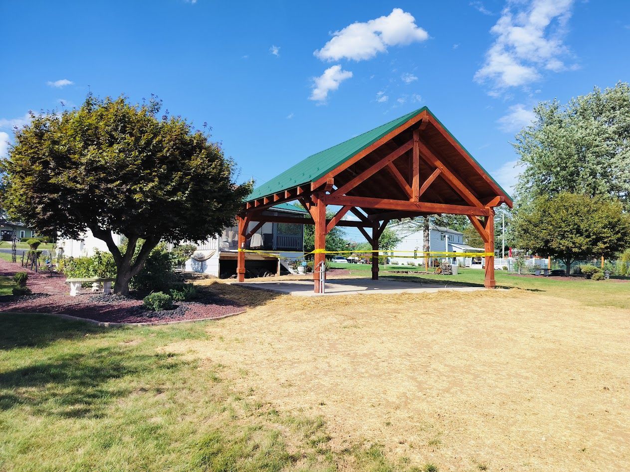 A wooden pavilion with a green roof stands on a lawn next to a tree under a blue sky.