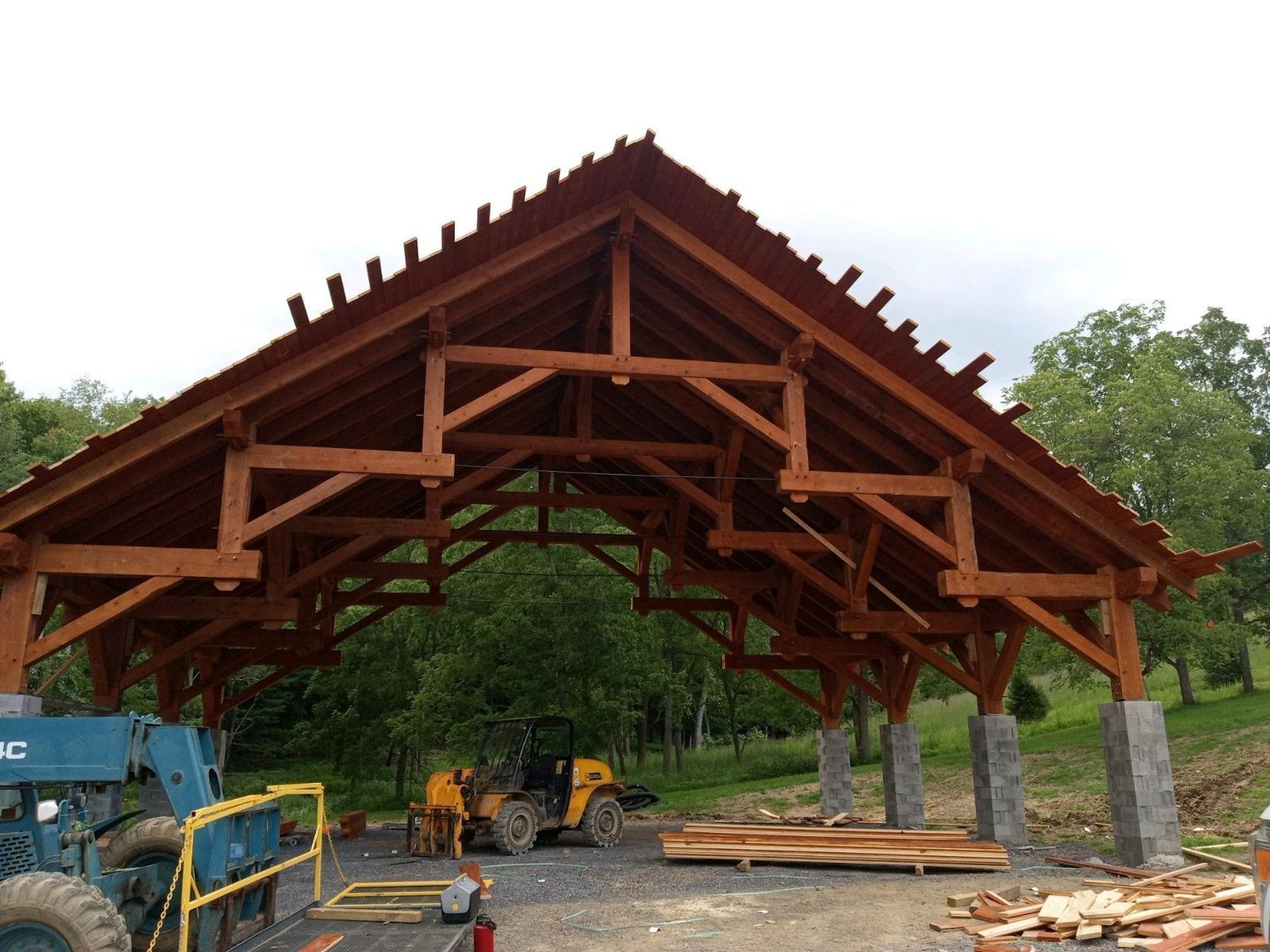 An unfinished timber frame pavilion under construction, featuring heavy wooden trusses supported by stone-clad columns.