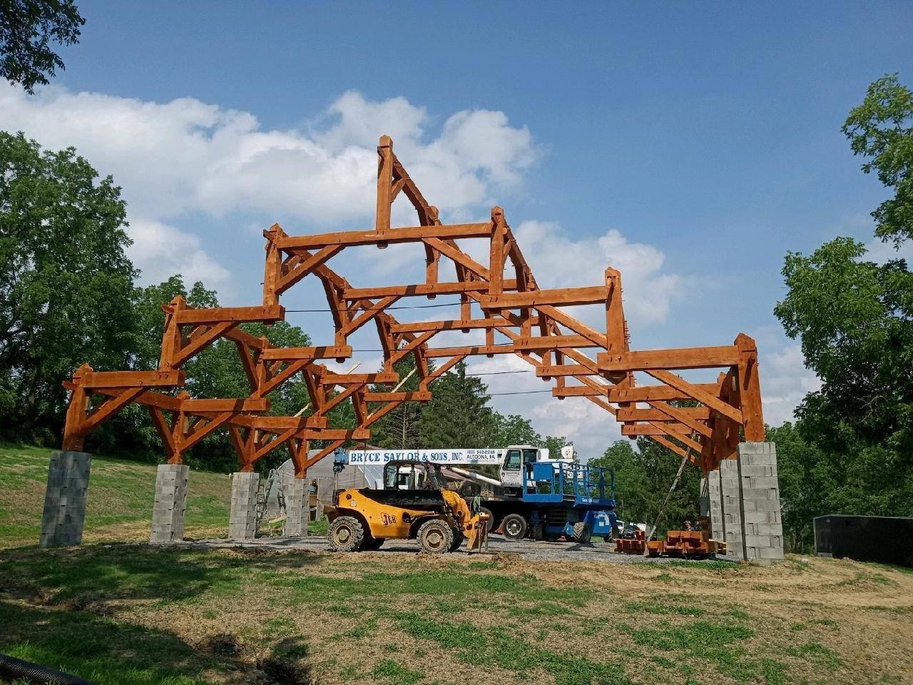 A large timber-frame structure under construction in an open, grassy field with a yellow telehandler vehicle nearby.