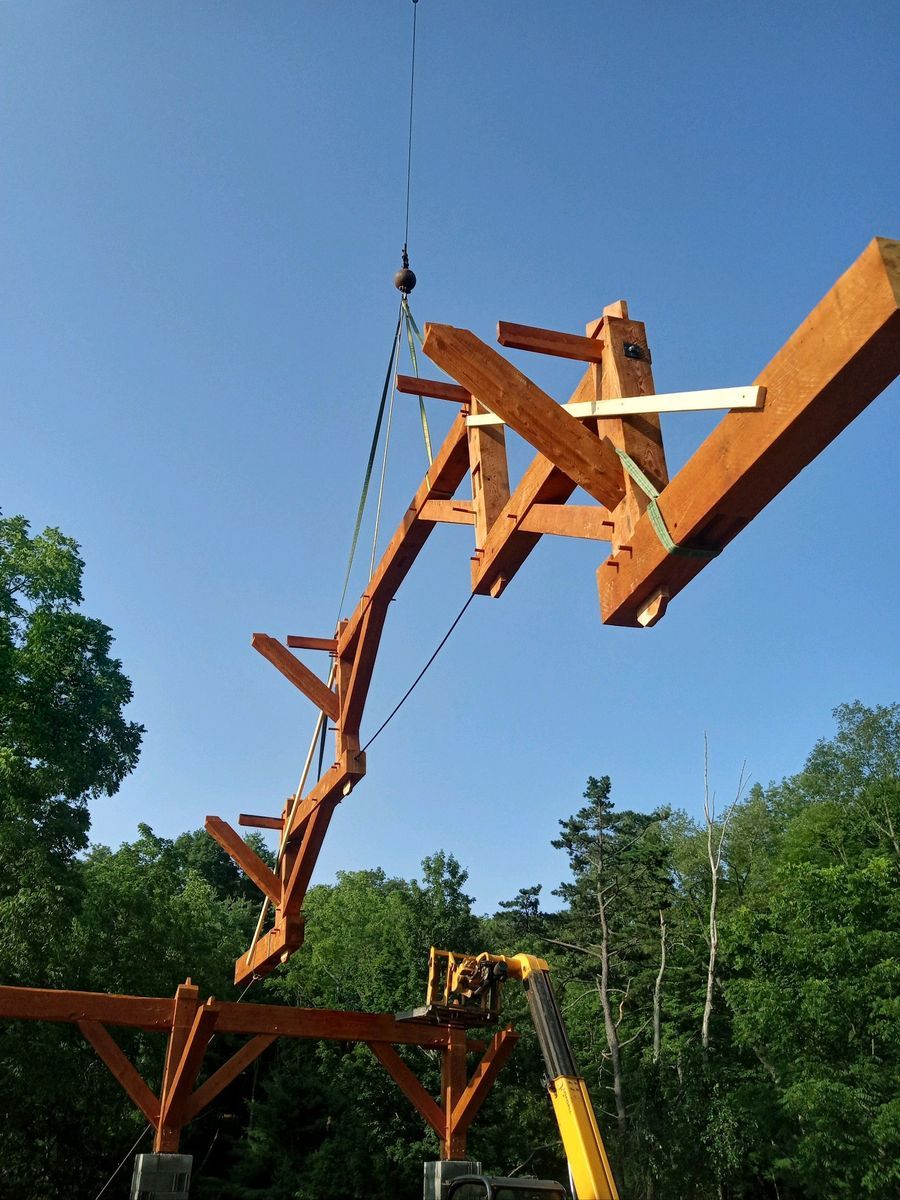 A crane lifts a large wooden timber-frame roof truss into position above a matching structure against a clear blue sky.