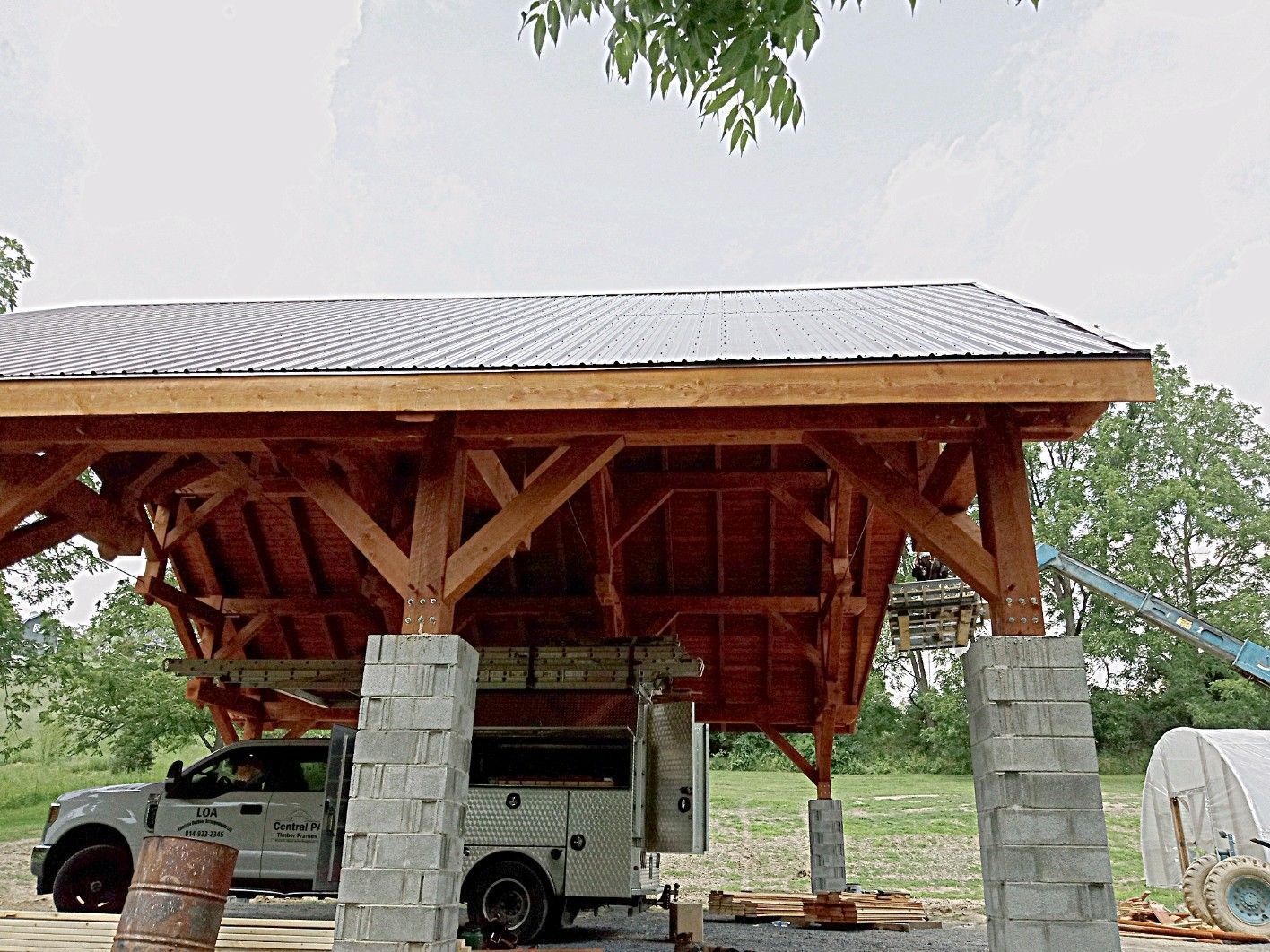 A large wooden timber-frame structure under construction, supported by concrete block pillars in a rural field.
