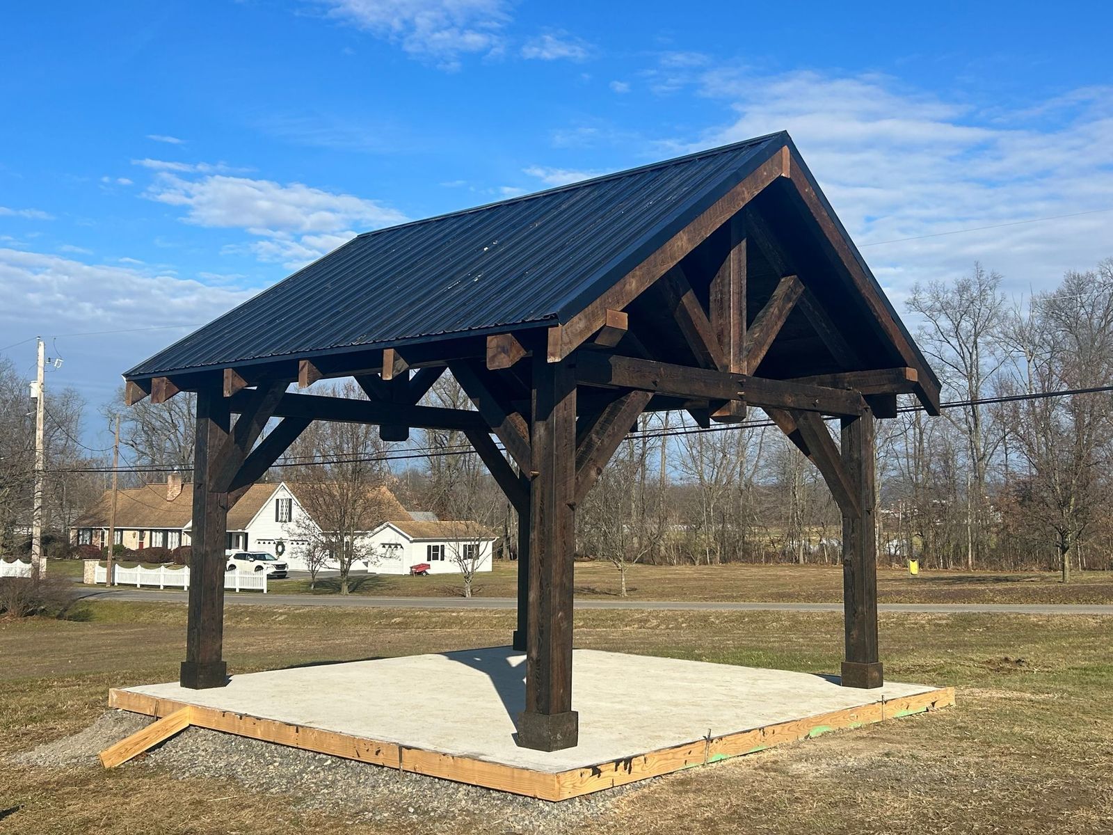 A dark-stained timber pavilion with a black metal gabled roof stands on a gravel foundation in a grassy field.