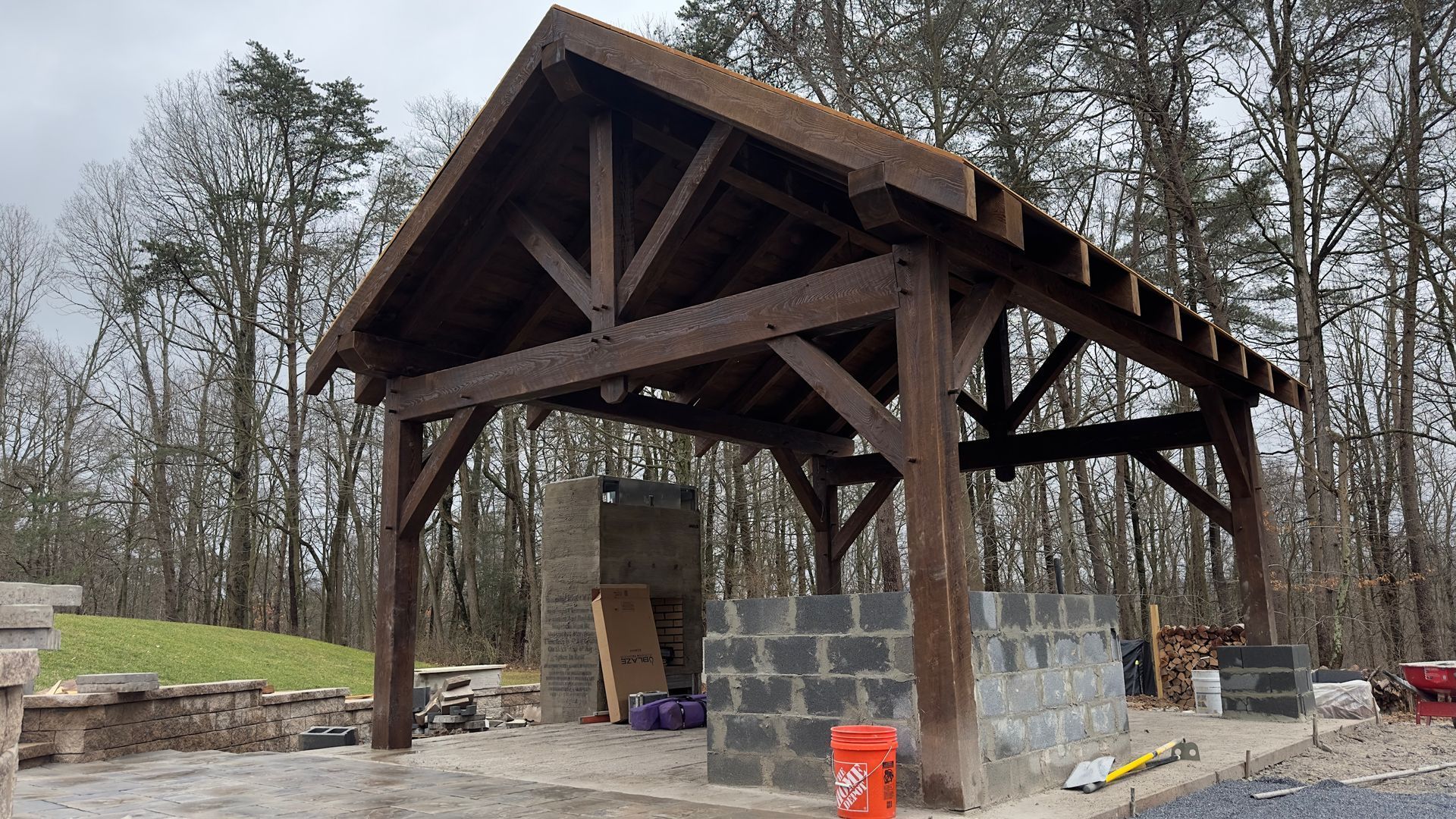 A timber frame pavilion under construction with a central cinder block structure, situated in a wooded area.