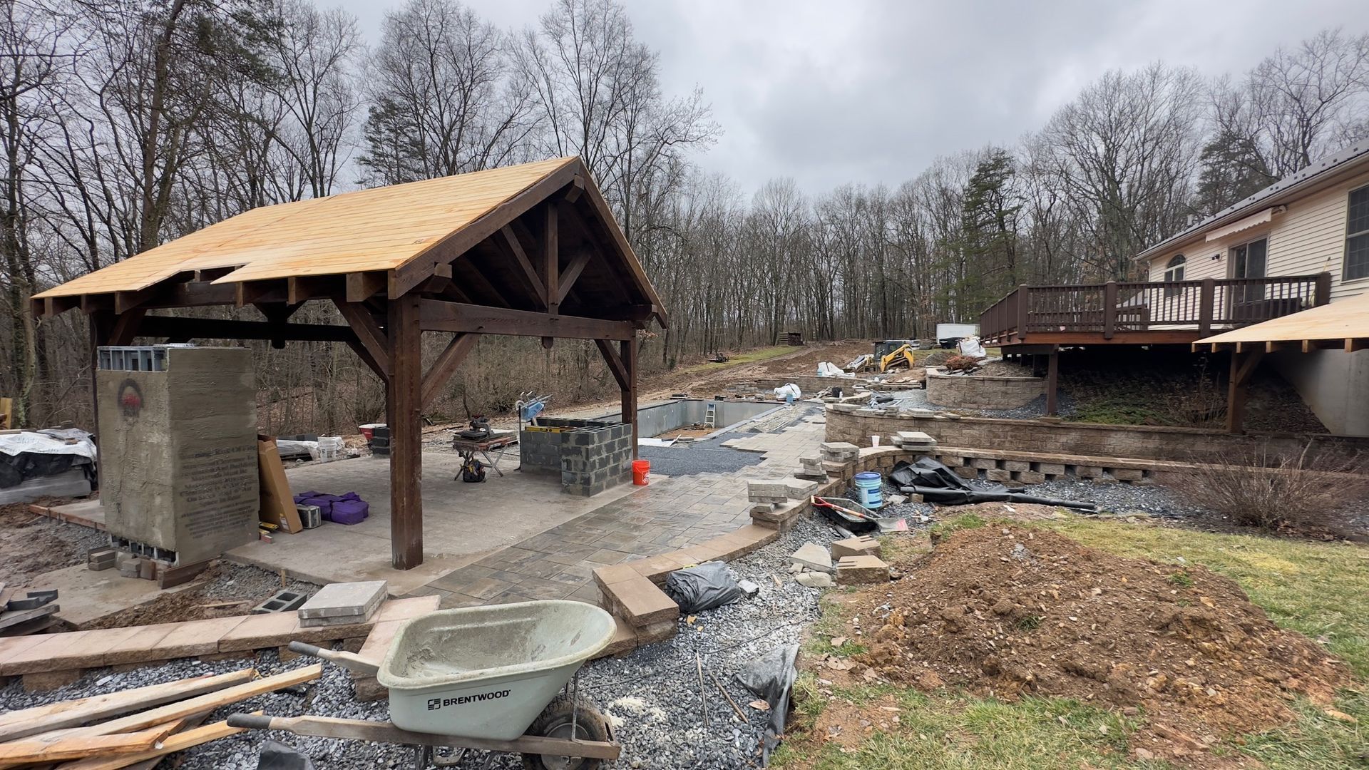A construction site featuring a wooden pavilion, a stone fireplace, a wheelbarrow, and a house with a deck in the background.