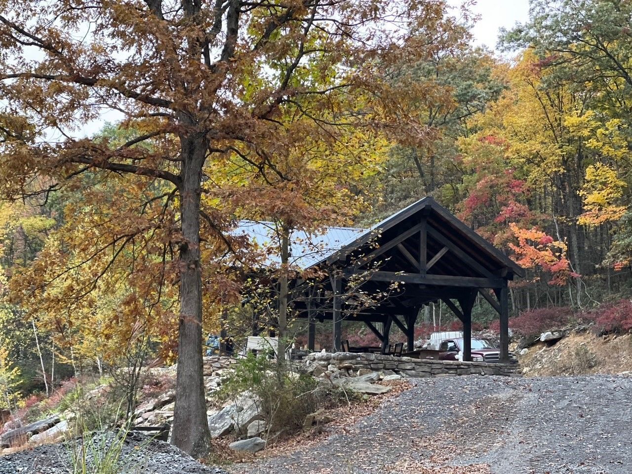 A wooden pavilion with a dark frame stands on a gravel path amidst a forest with colorful autumn foliage.