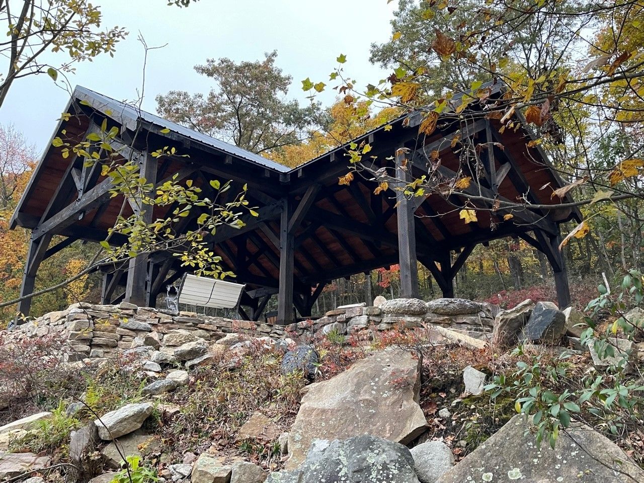 A rustic, wooden, double-gabled shelter stands on a rocky, tree-covered hillside during the autumn season.