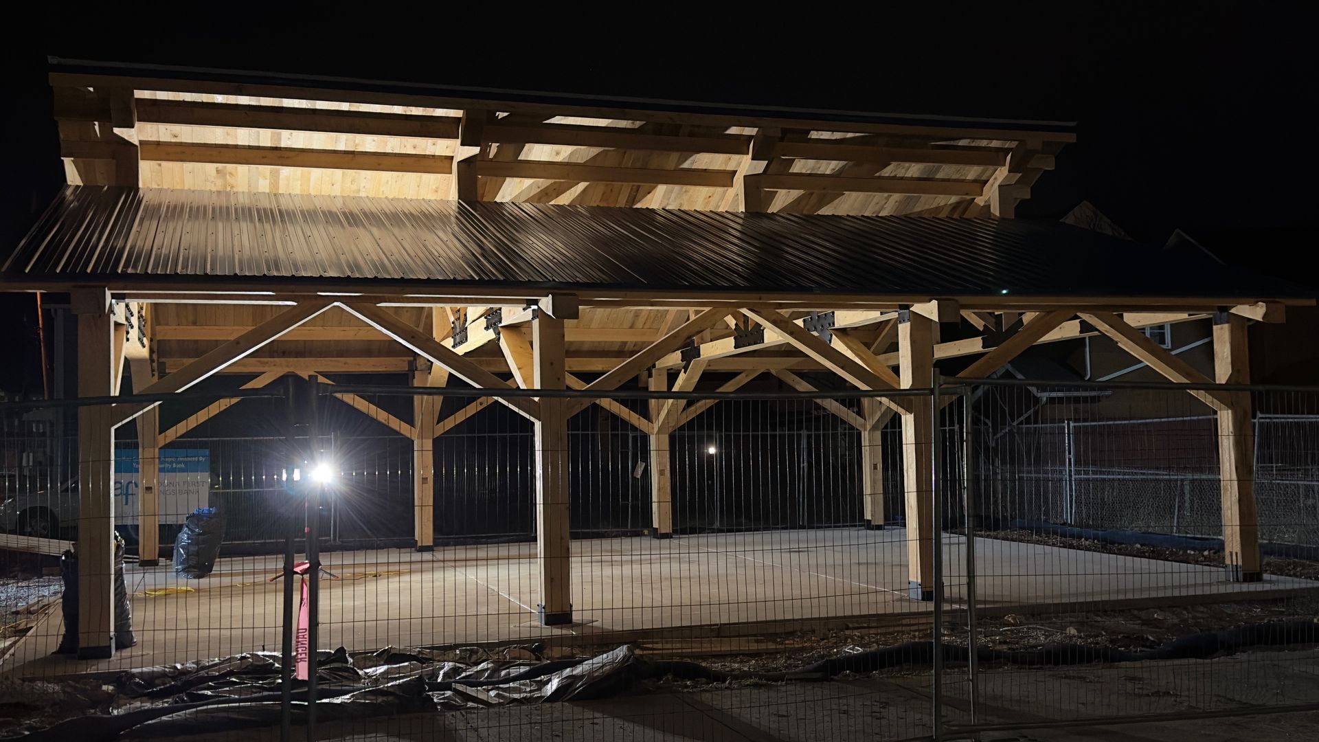 A timber-framed outdoor shelter with a metal roof is illuminated at night over a concrete pad with rebar sticking up.