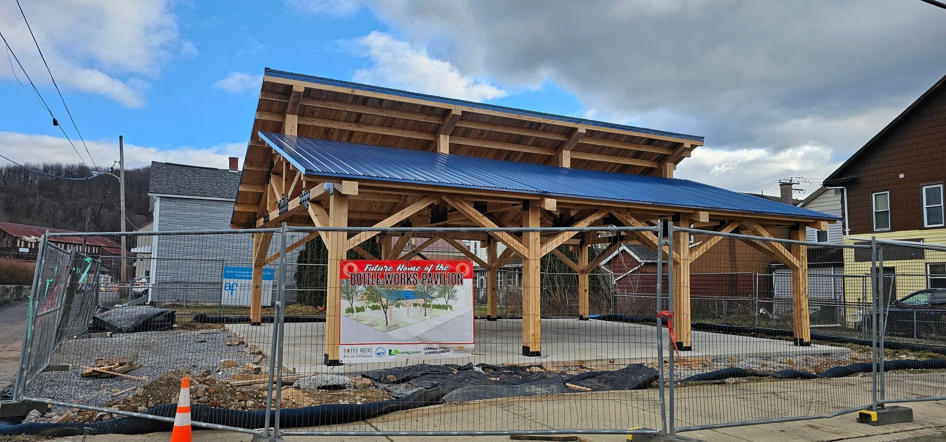 A timber-frame pavilion with a blue metal roof under construction, surrounded by temporary fencing on a concrete slab.