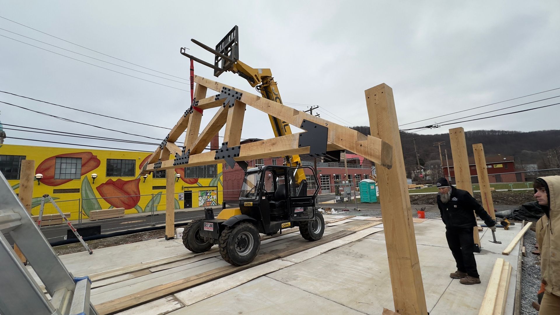 A telehandler lifts a large timber frame truss assembly onto vertical support beams at an outdoor construction site.