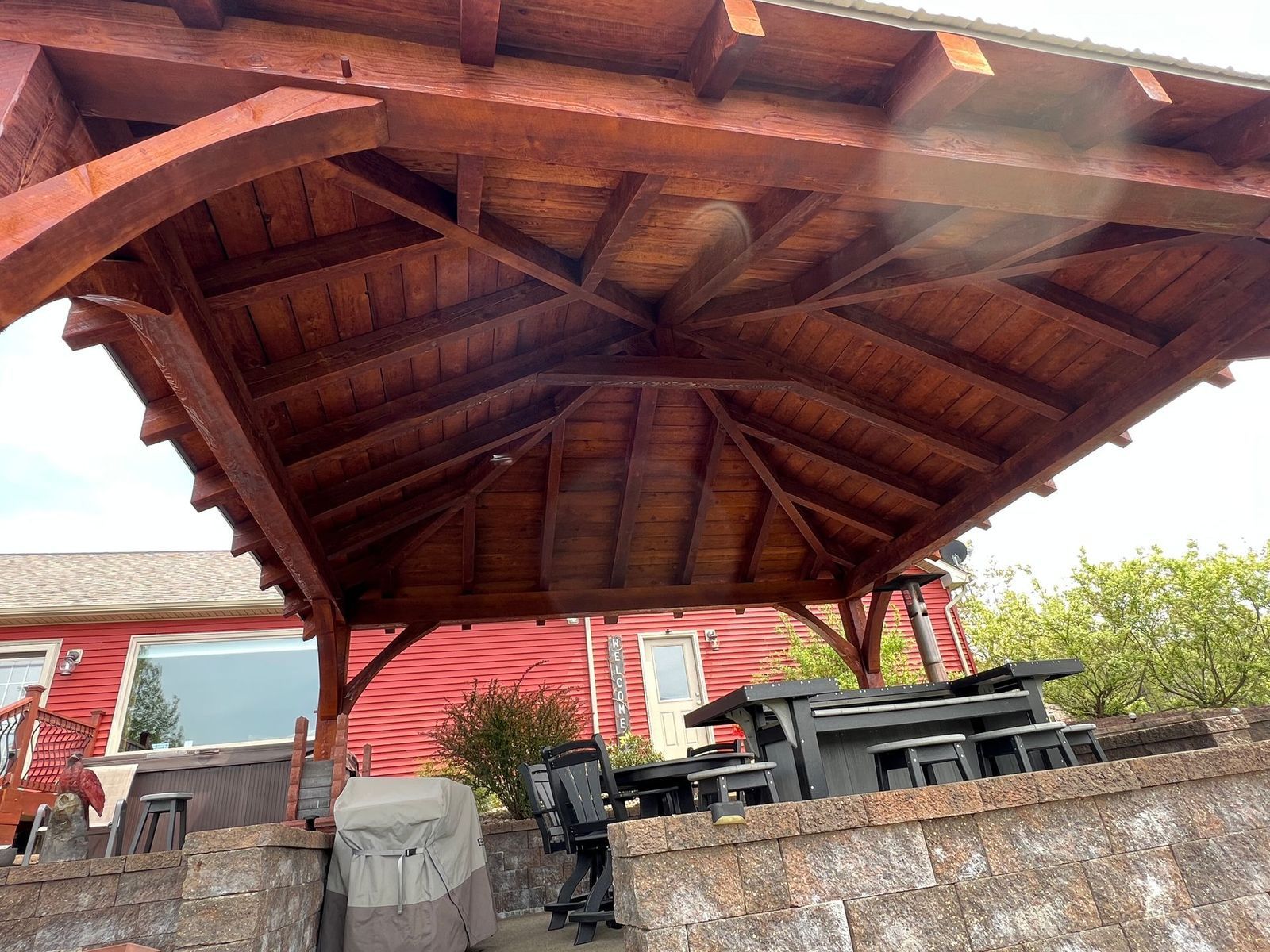 A dark wood gazebo structure over an outdoor patio with a dining set and grill against a red wall.