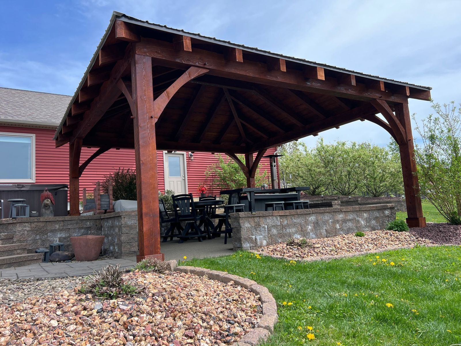 A wooden pavilion structure over a patio with outdoor furniture next to a red building.