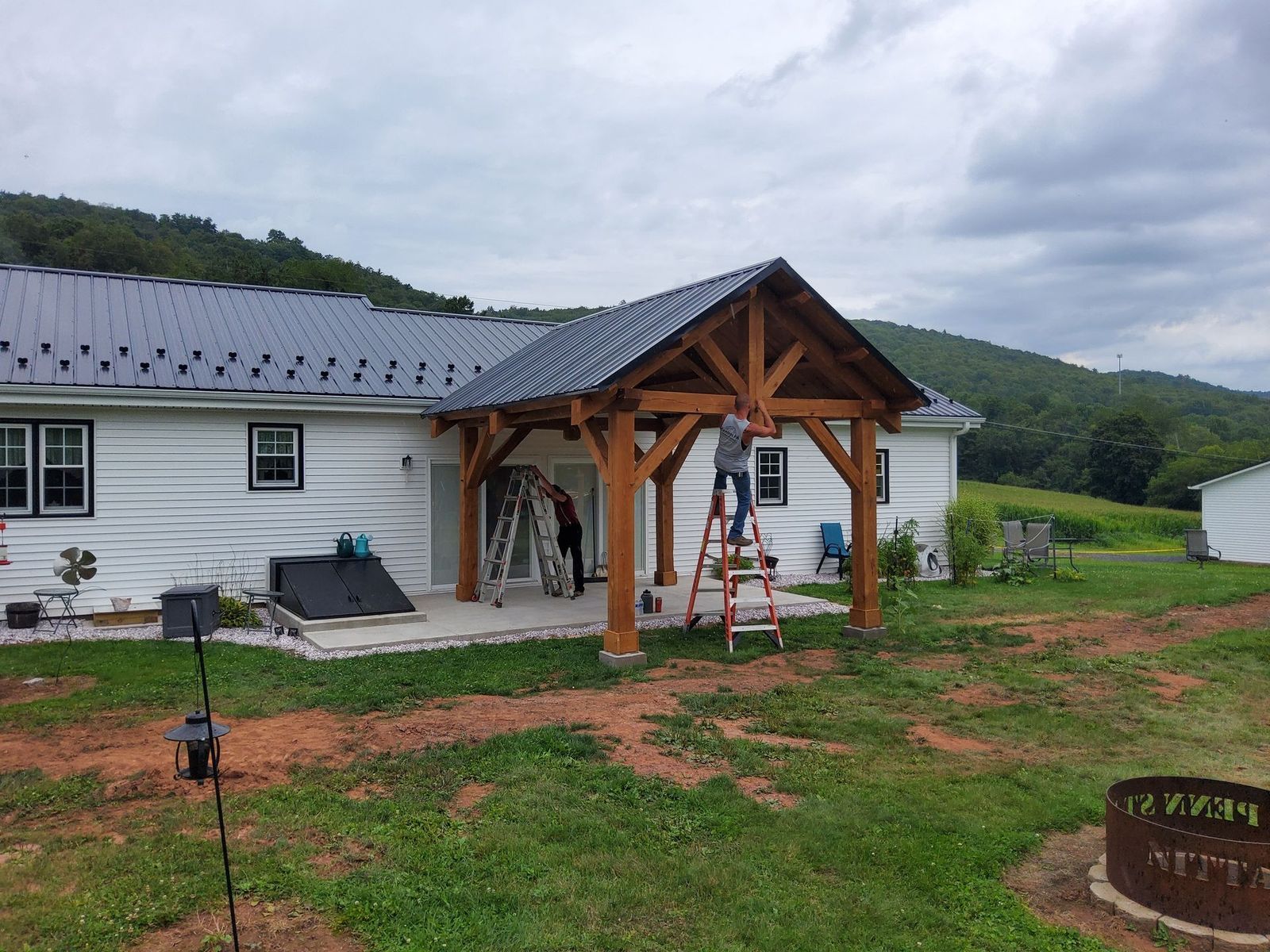 Workers construct a timber-frame porch attached to a white house with a metal roof under a cloudy sky.