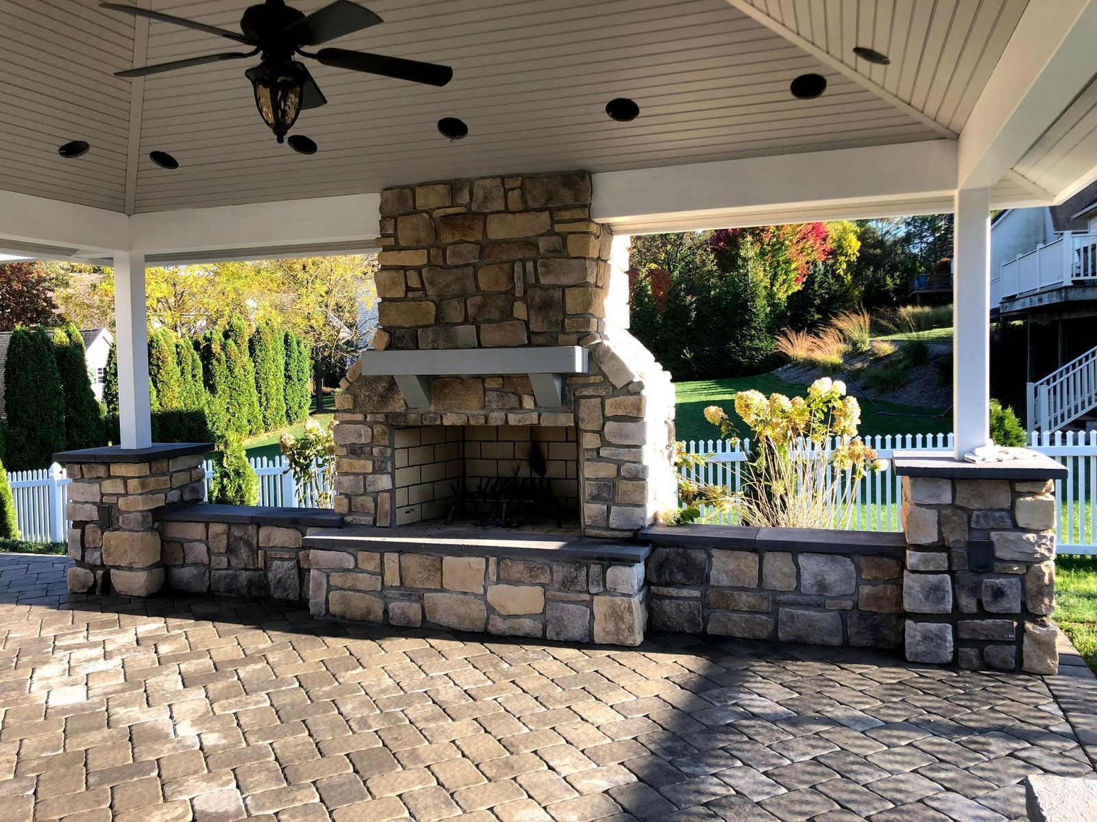 An outdoor stone fireplace and seating wall built on a patterned paver patio under a covered pavilion.