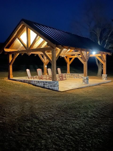Illuminated wooden pavilion with a metal roof and stone base, featuring four rocking chairs on a patio at dusk.