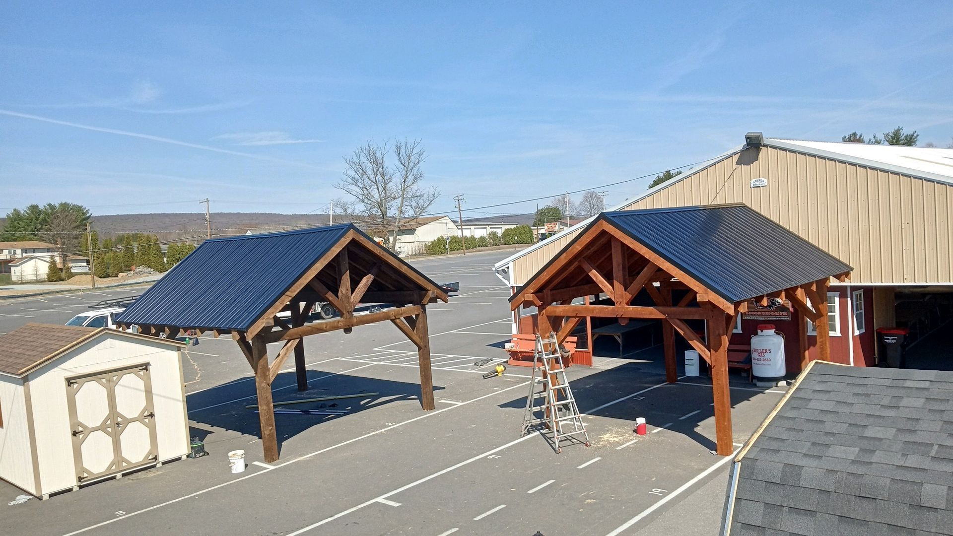 Two wooden pavilions with dark metal roofs stand in an outdoor parking lot next to a small shed and a larger building.
