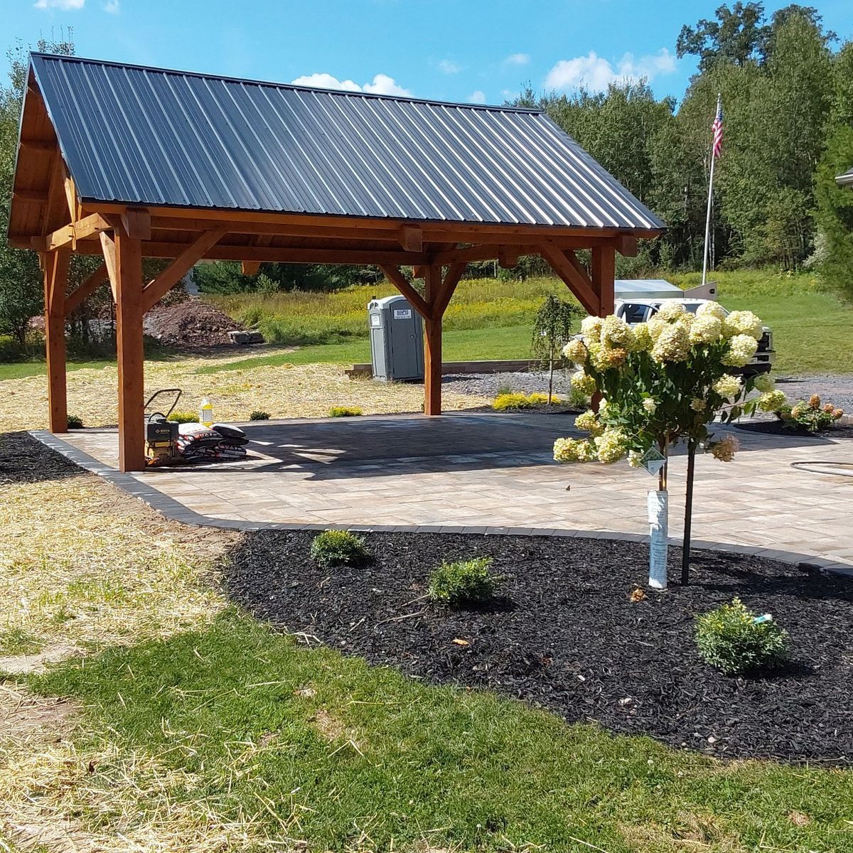 A wooden timber-frame pavilion with a black metal roof over a paved patio, near a flower bush in a mulched bed.