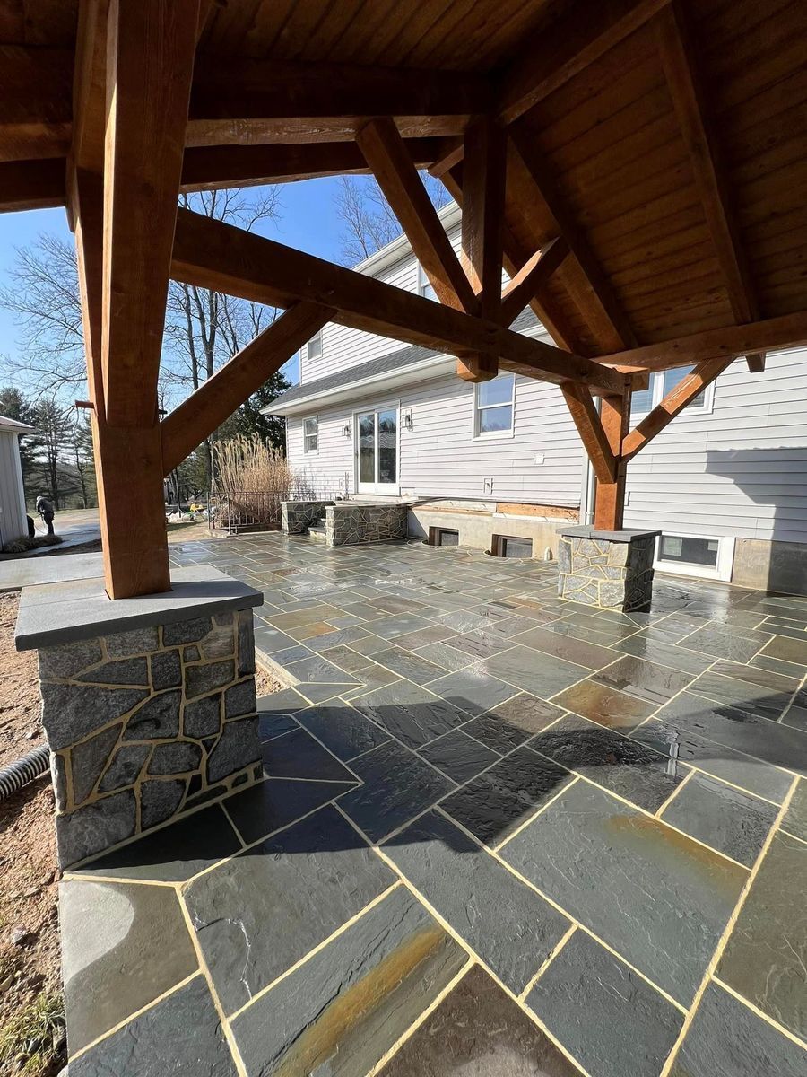 A timber-framed patio cover over a dark gray natural stone patio next to a house with light gray siding.