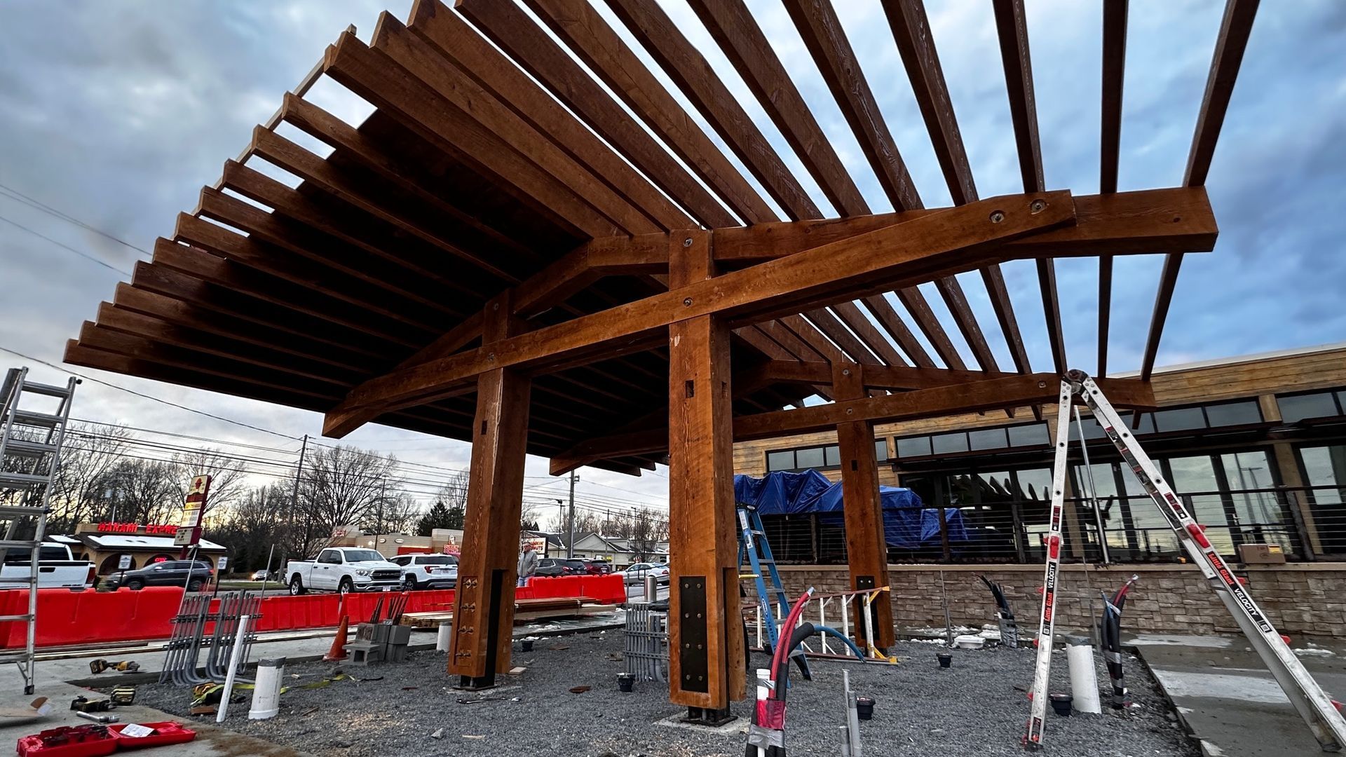 A wooden pergola structure under construction with a sloped roof over a gravel ground at a job site.