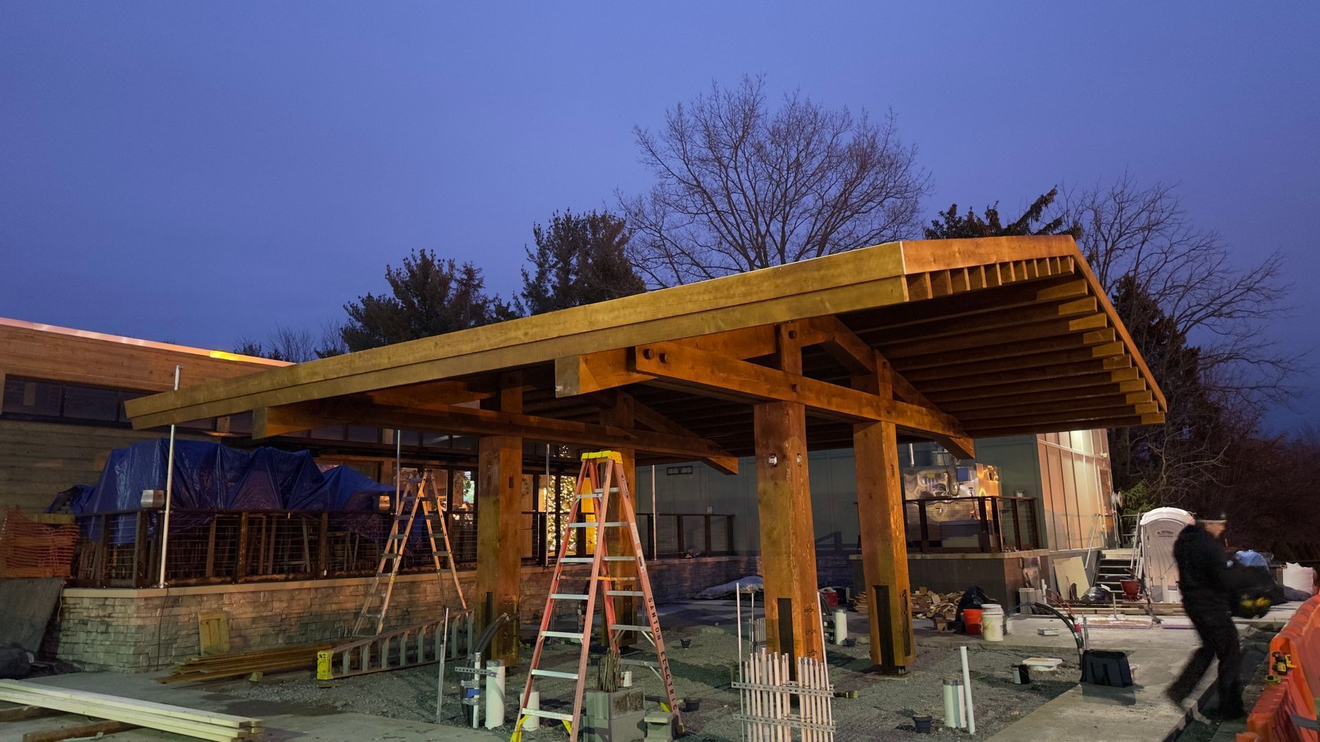 A construction crew works on a wooden timber-frame canopy structure at twilight, with scaffolding visible underneath.