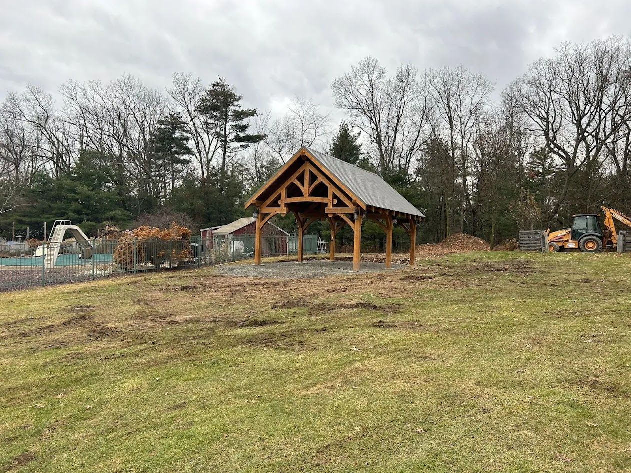 A wooden timber-frame pavilion stands in an open field, with construction equipment and a forested area in the background.