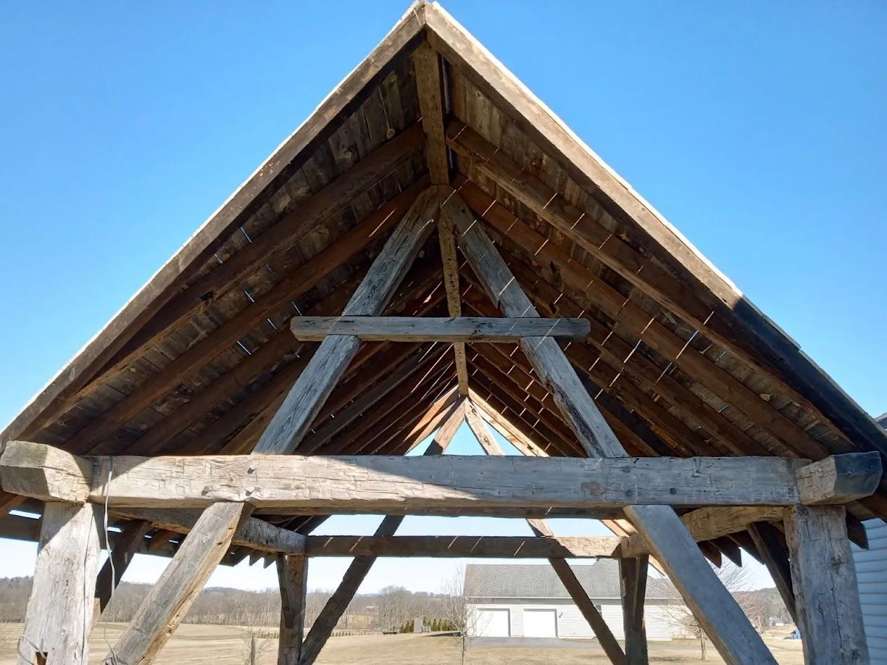 Low-angle view of an open-air timber frame structure with an exposed wooden roof skeleton against a clear blue sky.