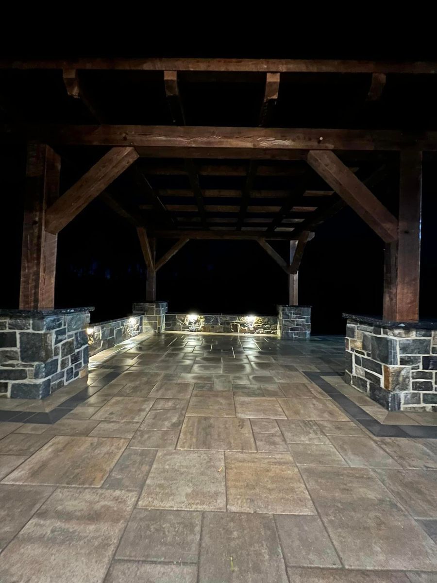 A wooden pergola covers a stone patio at night, illuminated by integrated wall and landscape lighting.
