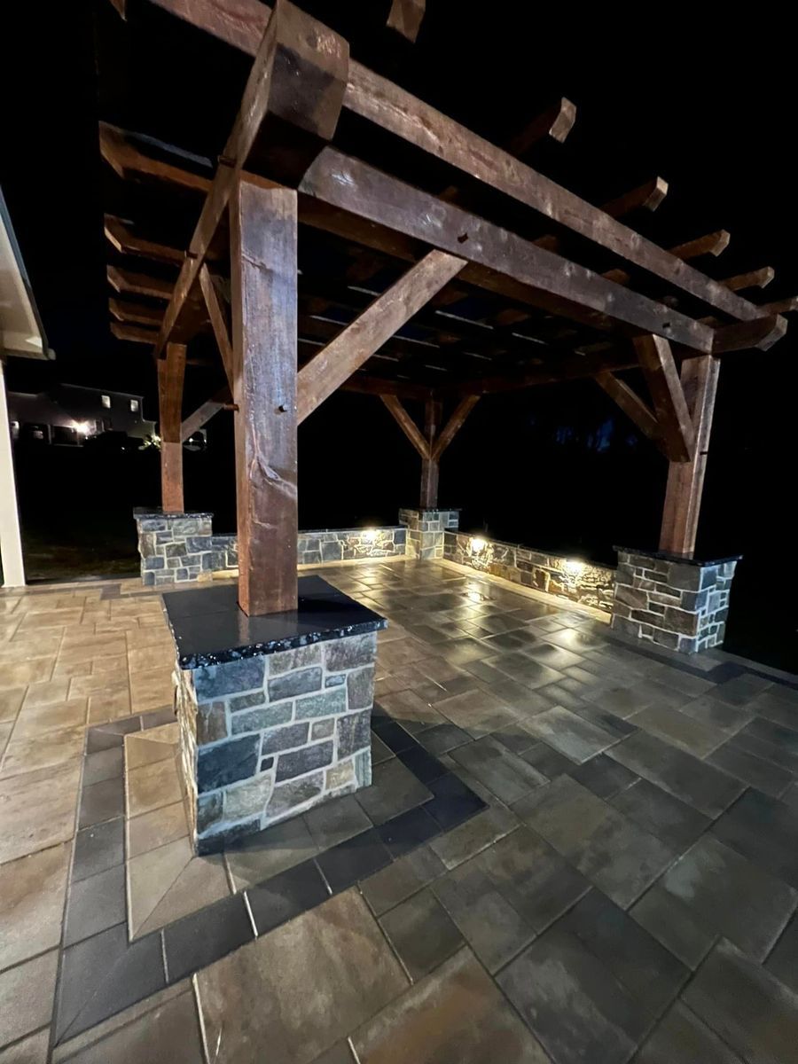 A wooden pergola on a stone patio at night, illuminated by integrated pillar and perimeter lighting.