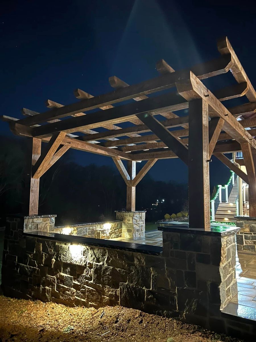 A wooden pergola over a stone patio with a low stone wall, illuminated by warm landscape lighting at night.