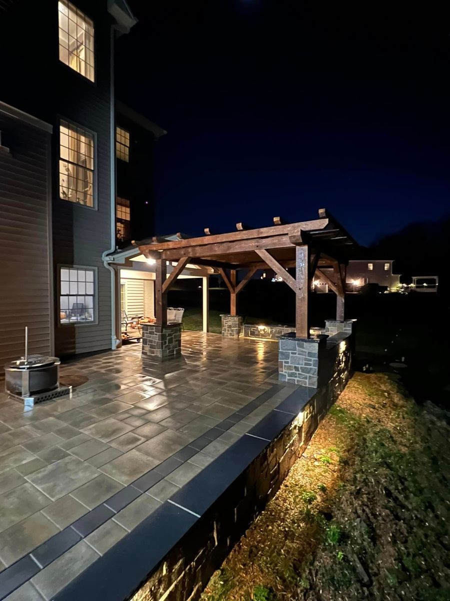 A backyard stone patio at night, featuring a lit wooden pergola and built-in stone wall seating.