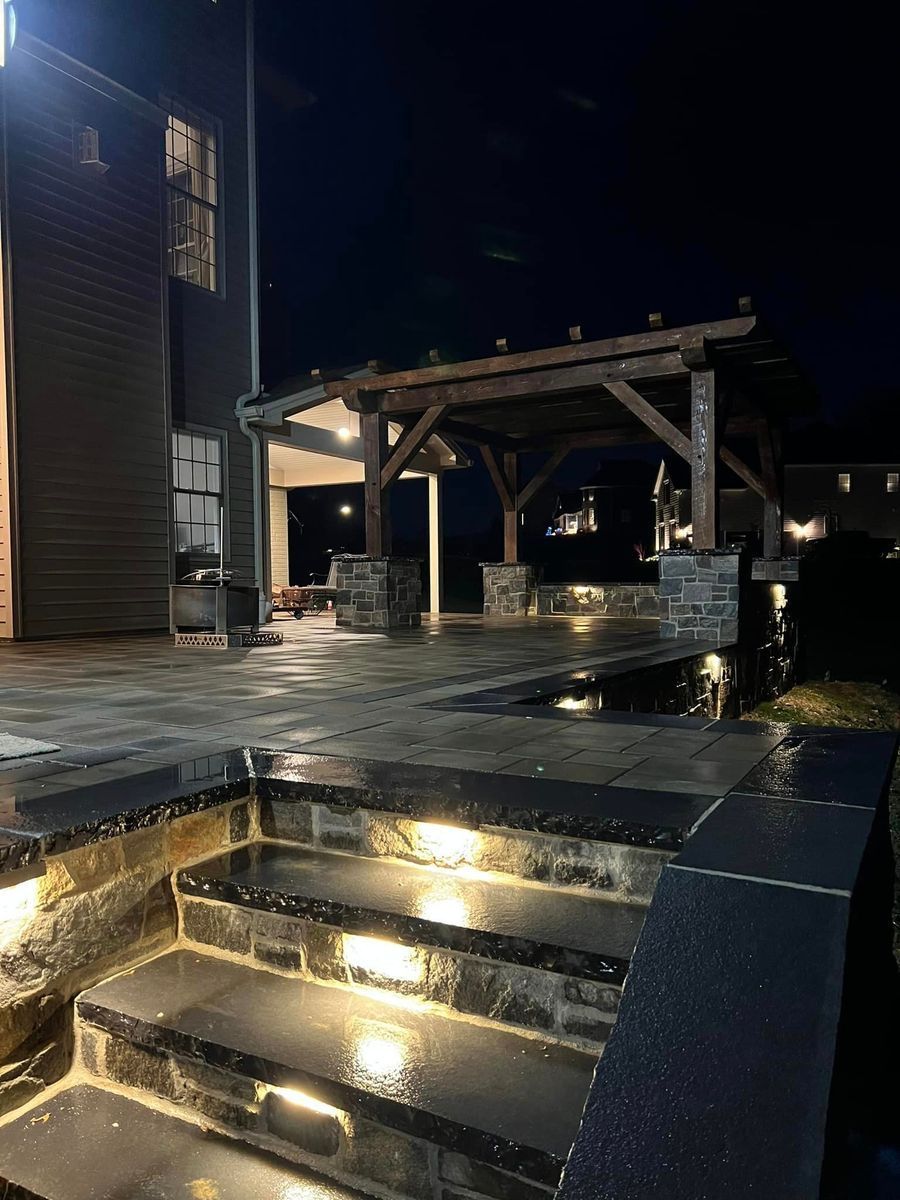 Central Timber Frame Supply's pergola. A stone patio at night featuring illuminated steps leading up to a wooden pergola.