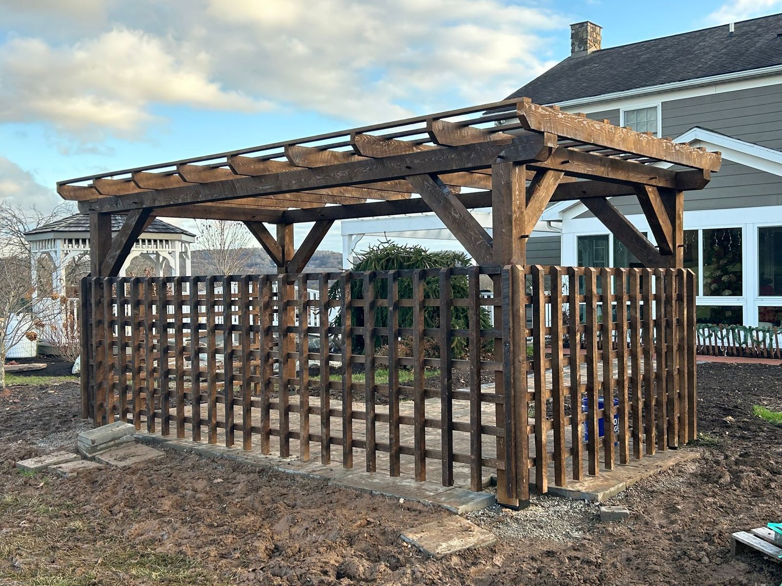 A dark wood pergola with a lattice fence sits on a stone patio in a yard beside a house under a cloudy sky. by CTFS.